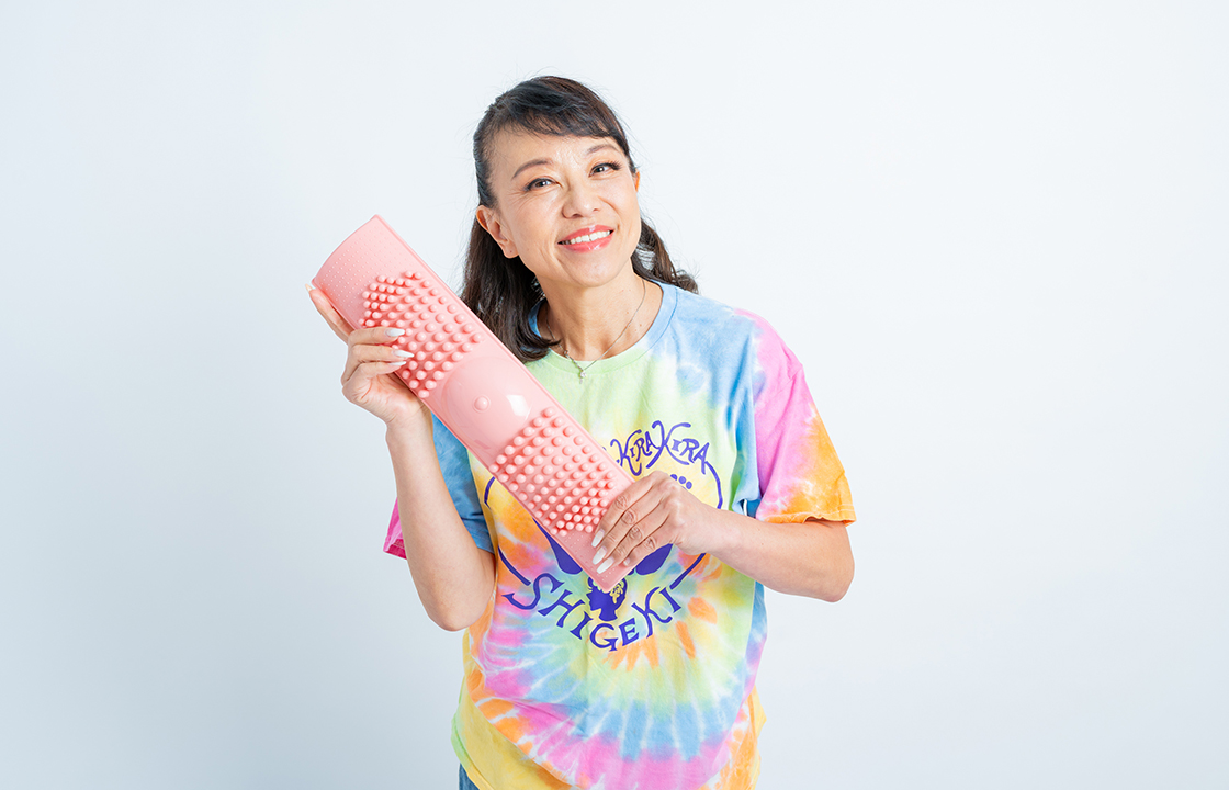 カラフルなTシャツの女性が足つぼグッズを手に笑顔で持っている様子