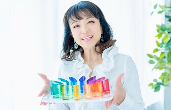 白いフリルのブラウスを着た女性が、カラフルなオブジェを手に持ち微笑んでいる様子