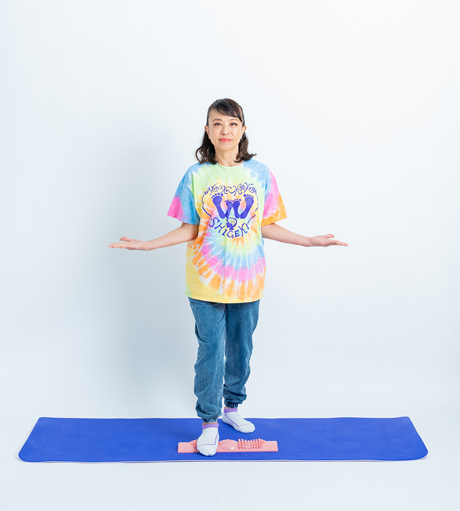 カラフルなTシャツを着た女性が足つぼグッズに乗って両手を広げている様子